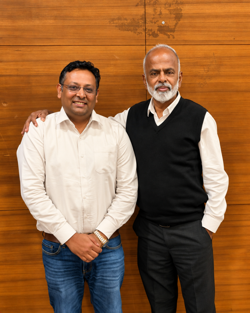 Two men posing together in front of a wooden wall, wearing formal and smart-casual outfits.